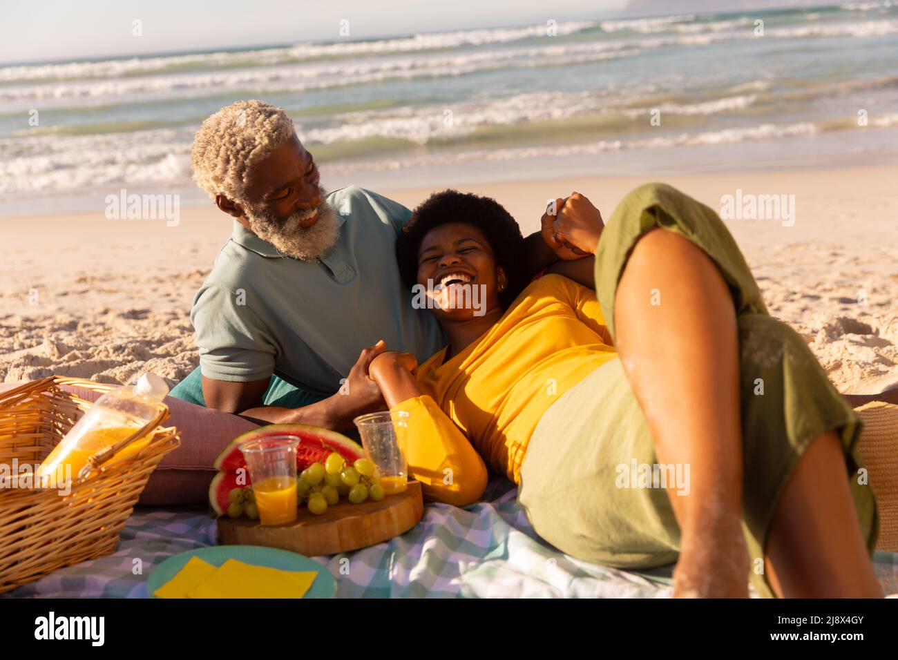 Cheerful african american mature woman lying by food and drink on senior man's lap against sea Stock Photo