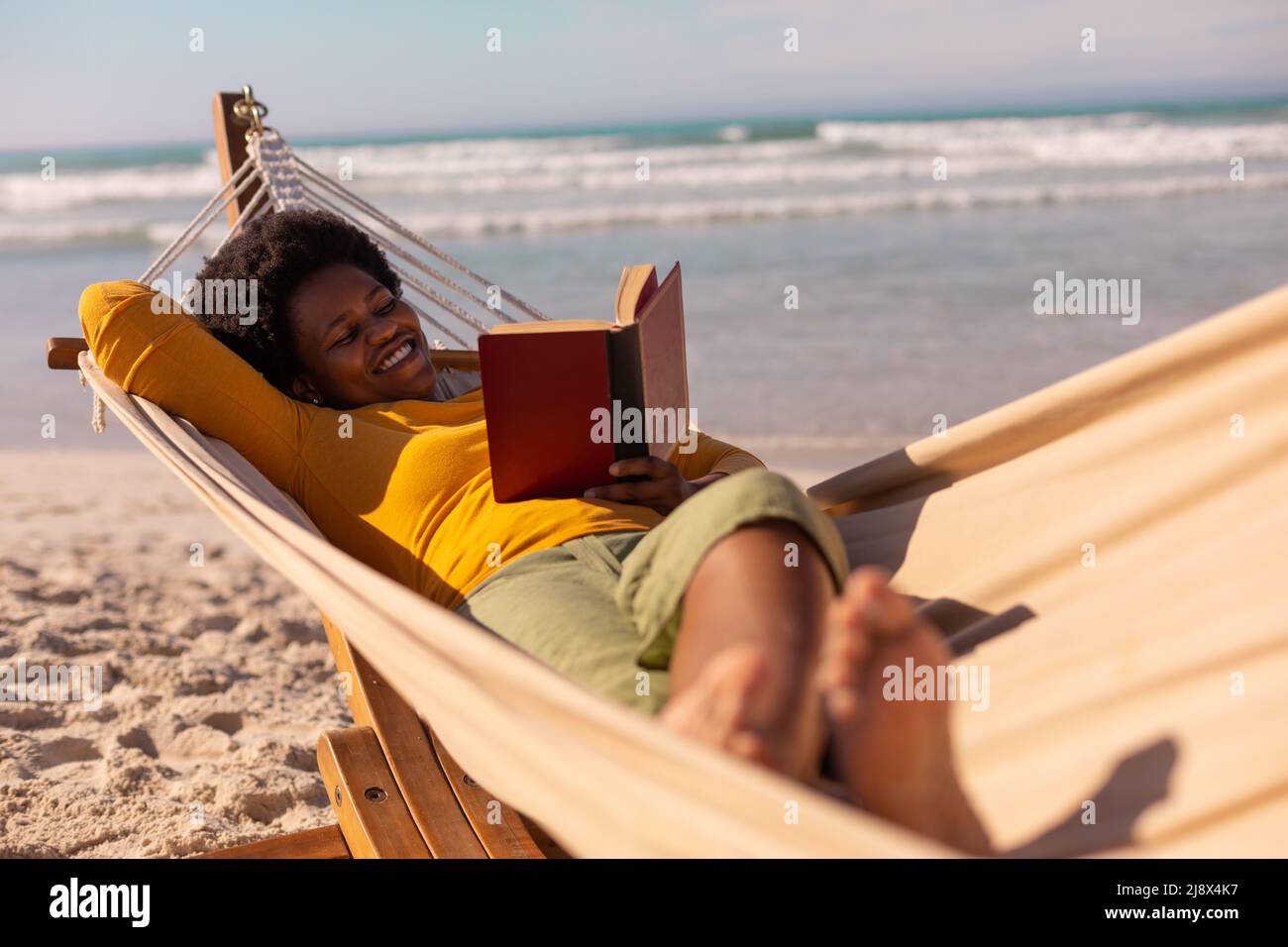 Smiling african american mature woman reading book while lying on hammock against sea in summer Stock Photo