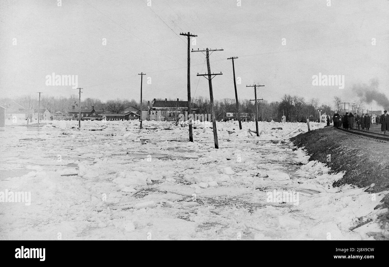 March 1918 Flood, Belleville, Ontario. Looking across the ice of the flooded Moira River to the east side. The big stone and brick building in the middle of the photo, was once called the Commercial Hotel and to the south of it is the Simpson/Wallbridge House. On the right of the photo is the Canadian Pacific Railway as it reaches the flooded west side of the river.  People are walking along the railroad right of way  ca.  1918 Stock Photo