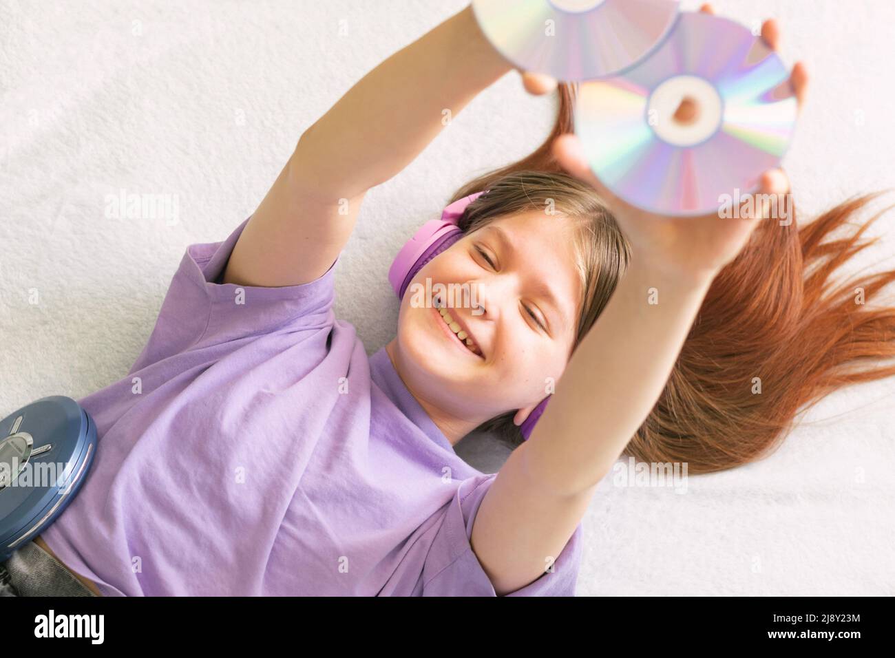 A happy teenage girl in a lilac T-shirt lies on the bed headphones and listens to music on CD player Stock Photo