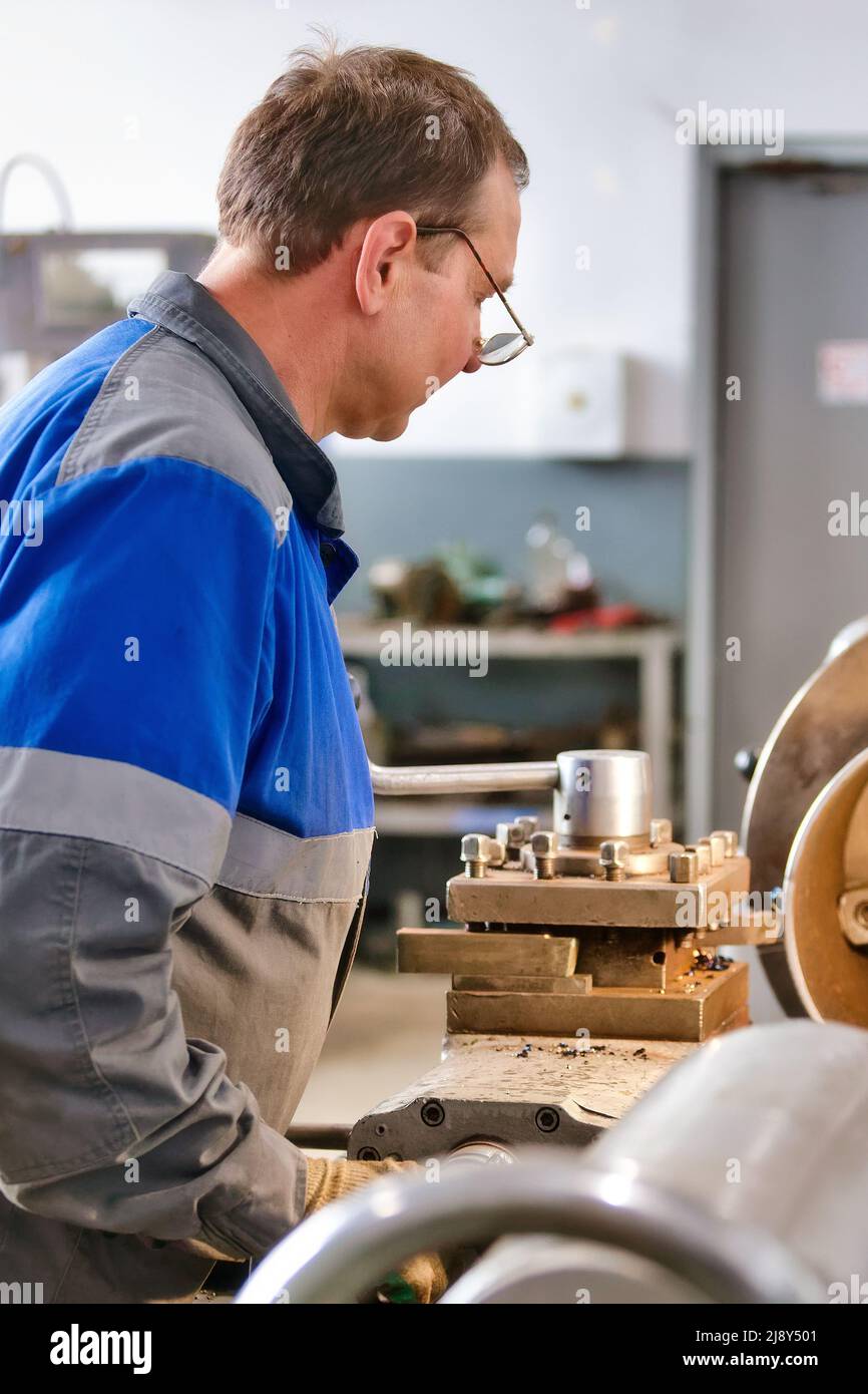 Elderly turner of 50-55 years old in overalls works at lathe in production shop. Processing of metal products. Real portrait of Caucasian worker. Production background. Stock Photo