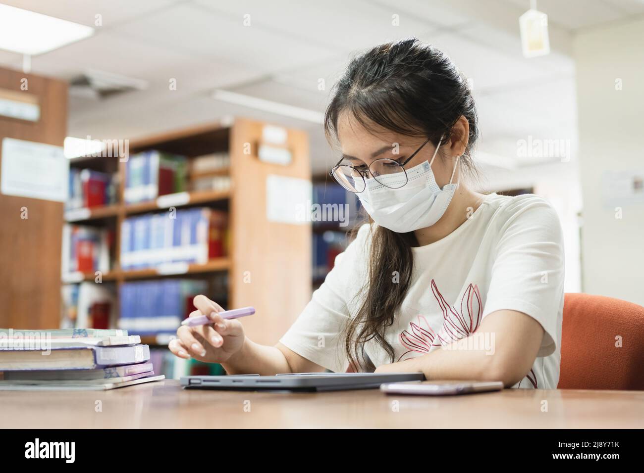 In the library, an Asian woman searches for information using a tablet and a book. Stock Photo
