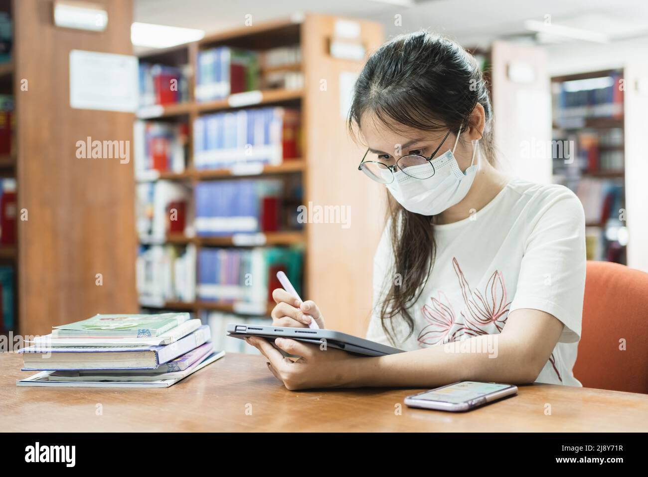 In the library, an Asian woman searches for information using a tablet and a book. Stock Photo