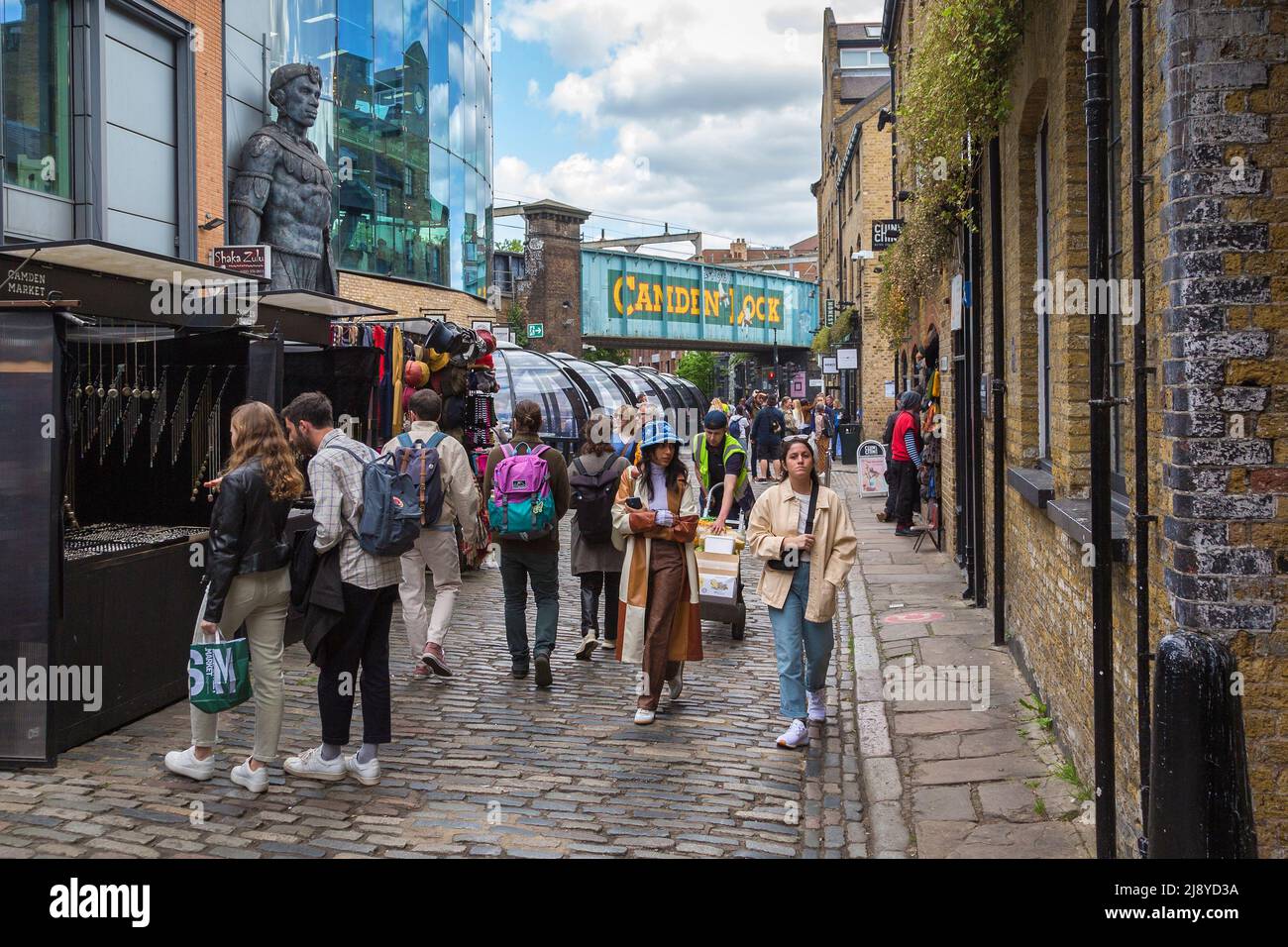A busy street filled with shoppers and tourists, with the iconic Camden Lock railway bridge in the background. Stock Photo