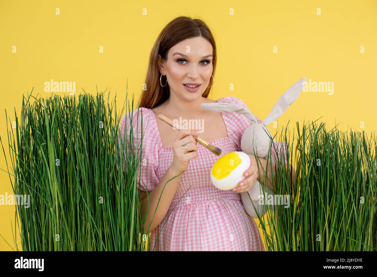 Woman in late pregnancy on grass decoration paint egg with brush and hold soft rabbit toy, yellow background. Portrait of young pregnant lady Stock Photo