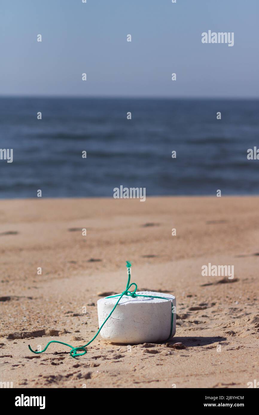 A shaped piece of white styrofoam with a green string around it standing on the sand near the ocean at the beach. Sea pollution concept Stock Photo