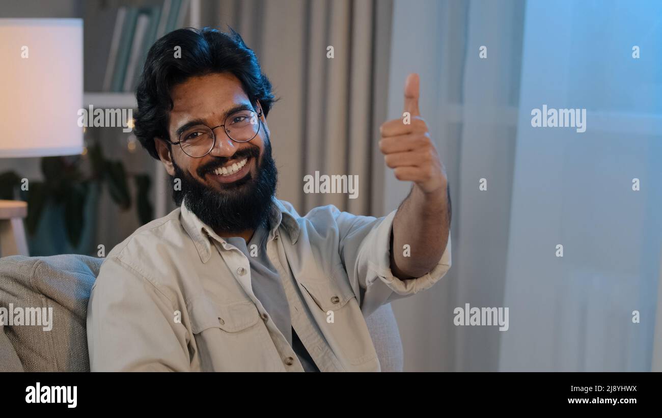 Portrait happy satisfied joyful arabic indian man bearded male in glasses sitting at home in new apartment on comfortable sofa looking at camera smili Stock Photo