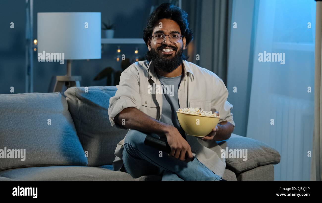 Arab adult bearded man in glasses indian male bachelor after work sits on  sofa in dark living room turns on TV with remote control choosing channel  wa Stock Photo - Alamy