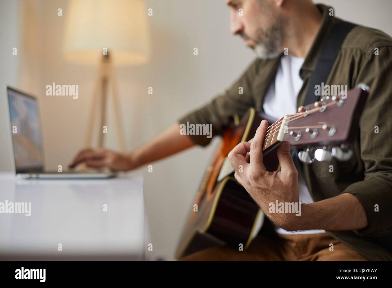 Man with acoustic guitar clamps desired chord while watching online lesson on laptop. Stock Photo