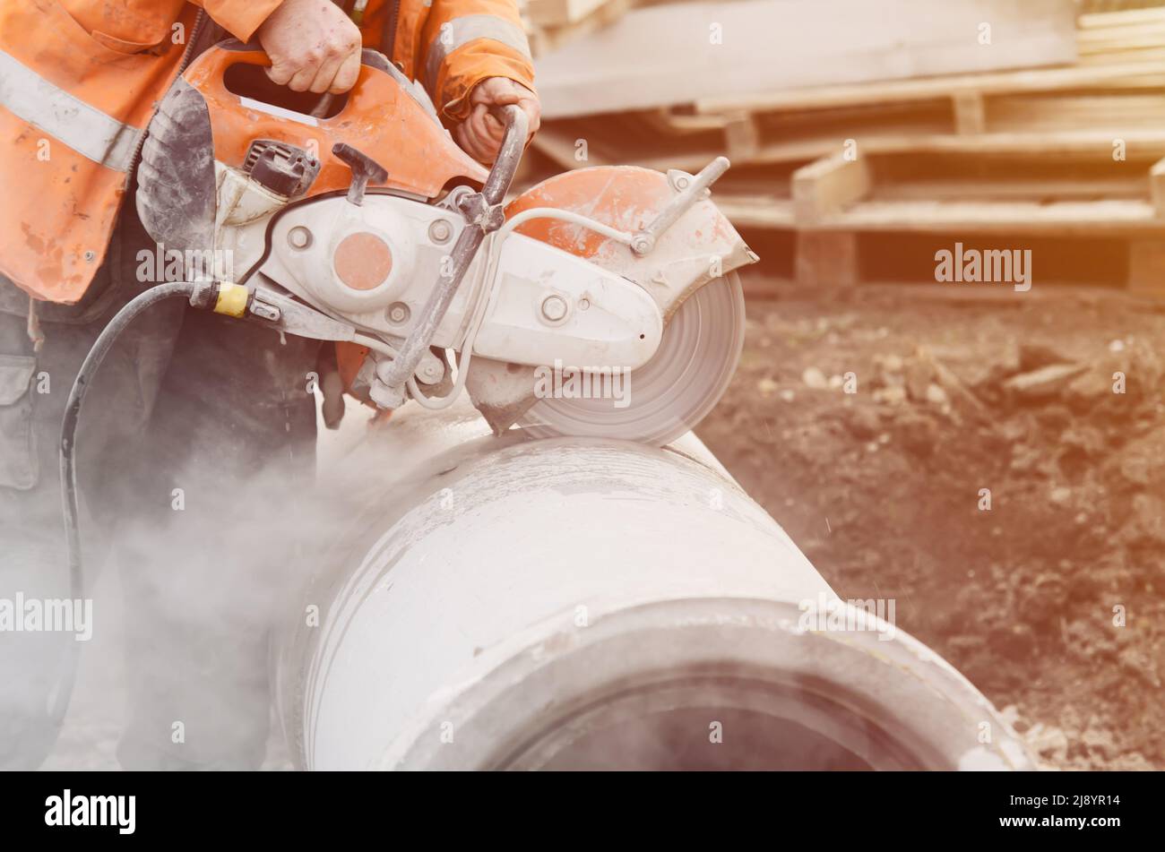A worker at the construction site cutting a concrete drainage pipe with a petrol concrete saw. Builder covered in a hazardous dust cloud as safety pro Stock Photo