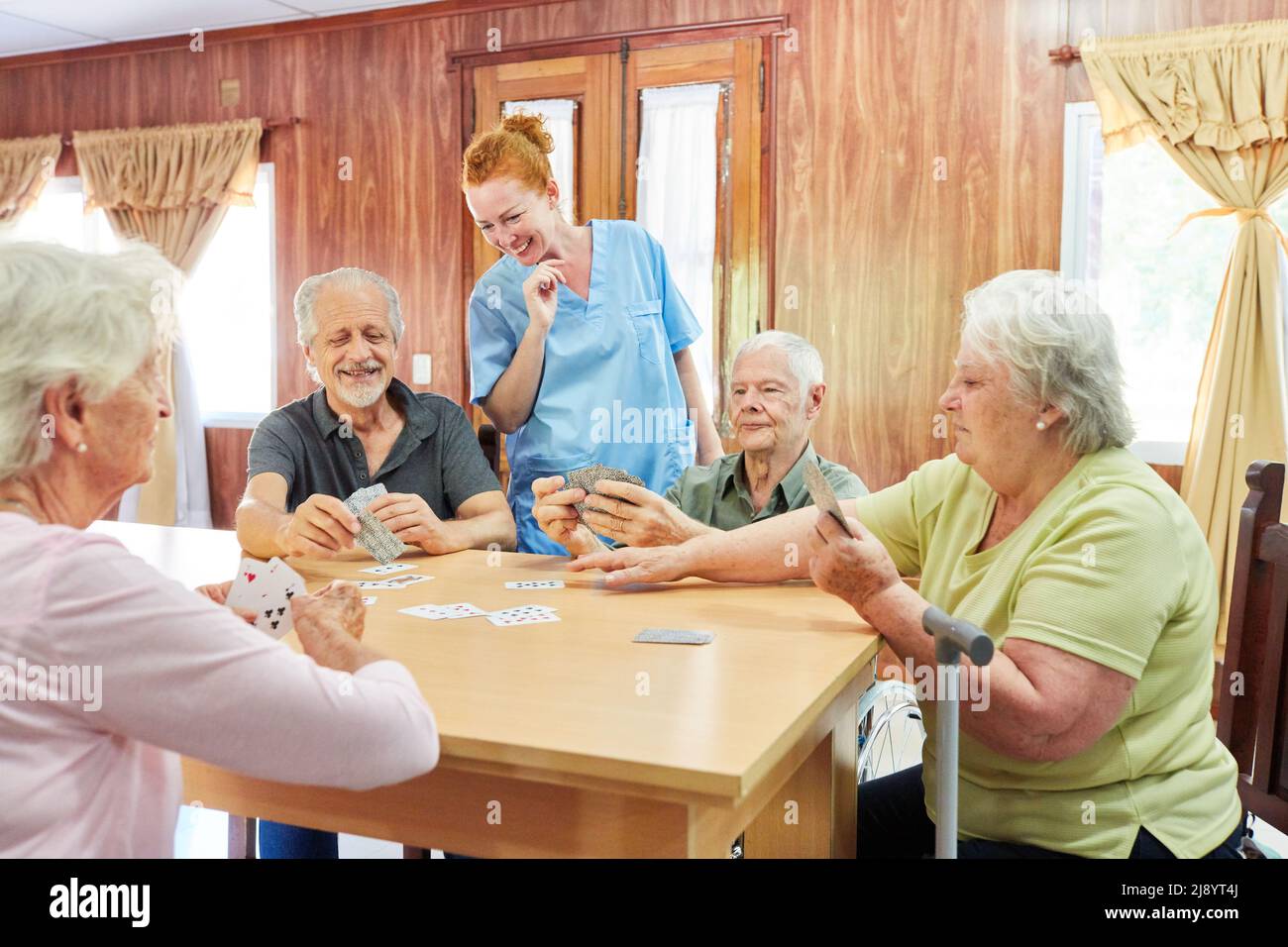 Group of seniors plays skat together at the table in the retirement home, supervised by a nurse Stock Photo