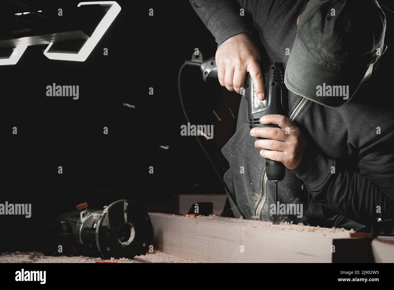 A man works with a drill in his workshop. Carpenter drills with a hand power tool in a dark room with directional light Stock Photo