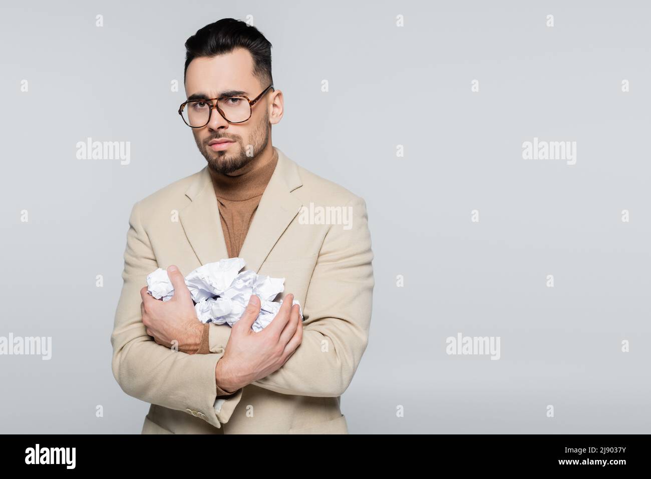 upset critic in eyeglasses looking at camera while holding crumpled paper isolated on grey Stock Photo