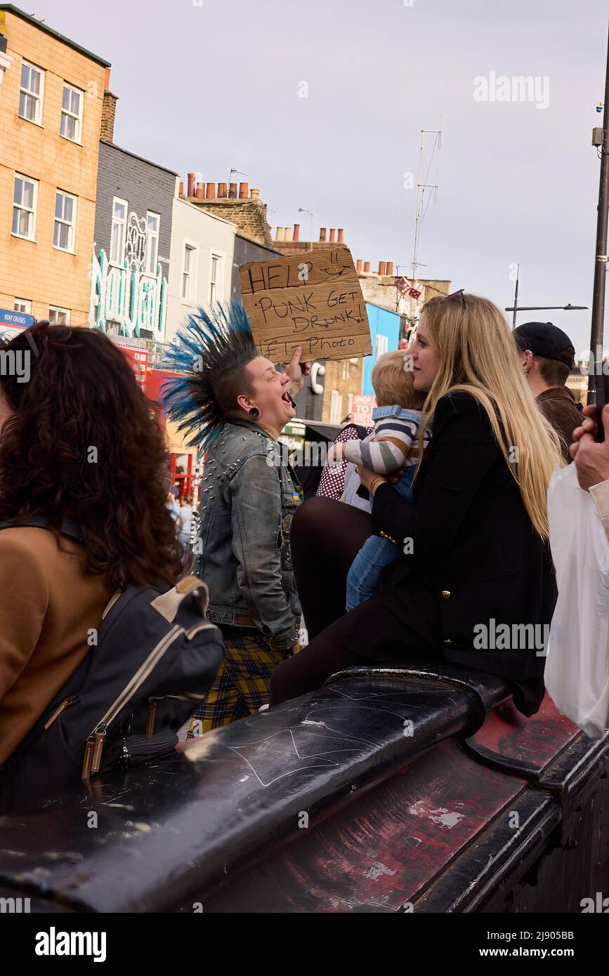 London punks with letter jacket and blue colored Liberty Spikes Mohawks on the crowded street having a good time holds a poster, help punks get drunk Stock Photo