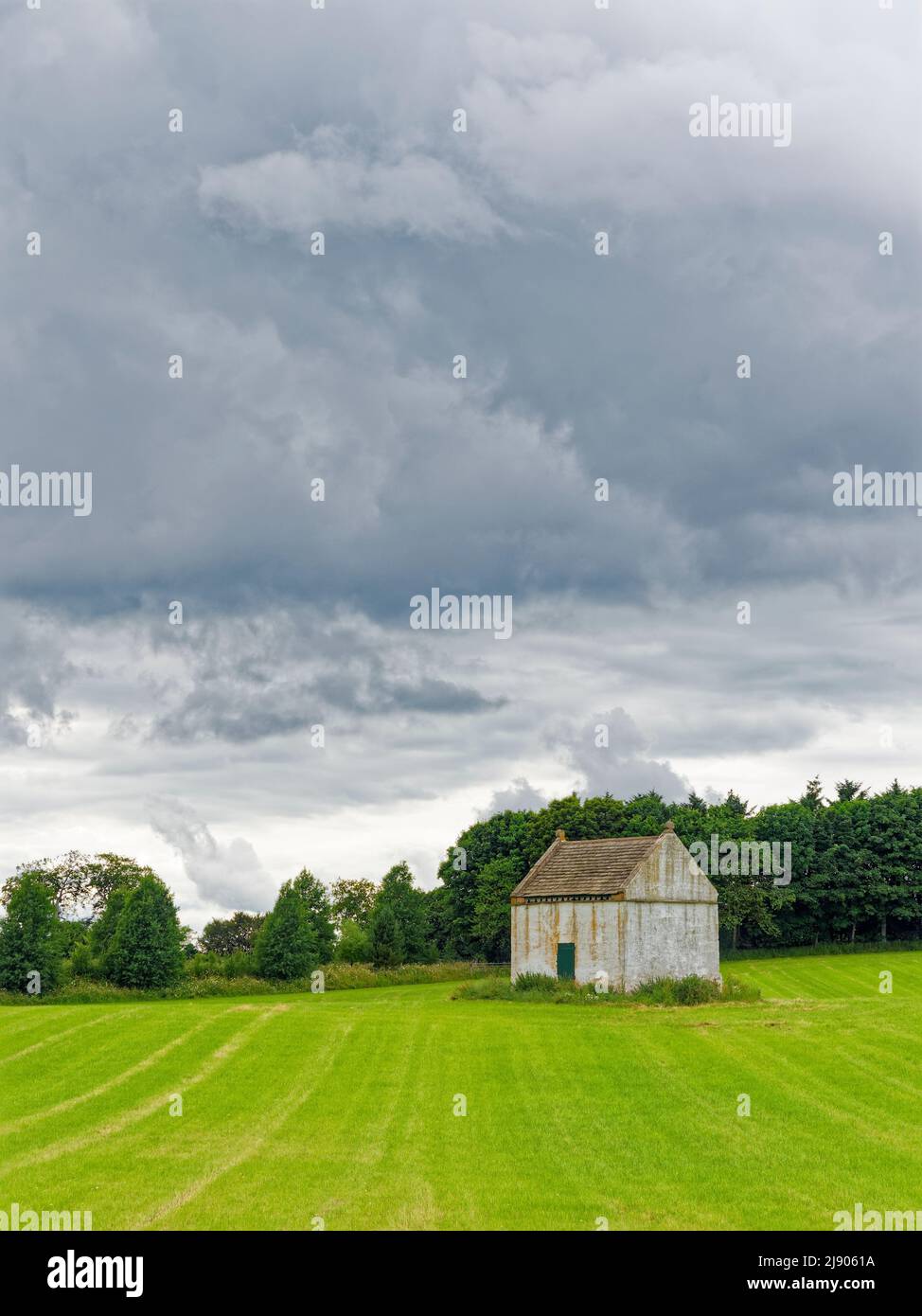 A pale white Old Doocot or Dovecote situated in a field of newly sown wheat with a mixed woodland behind and dark clouds above. Stock Photo