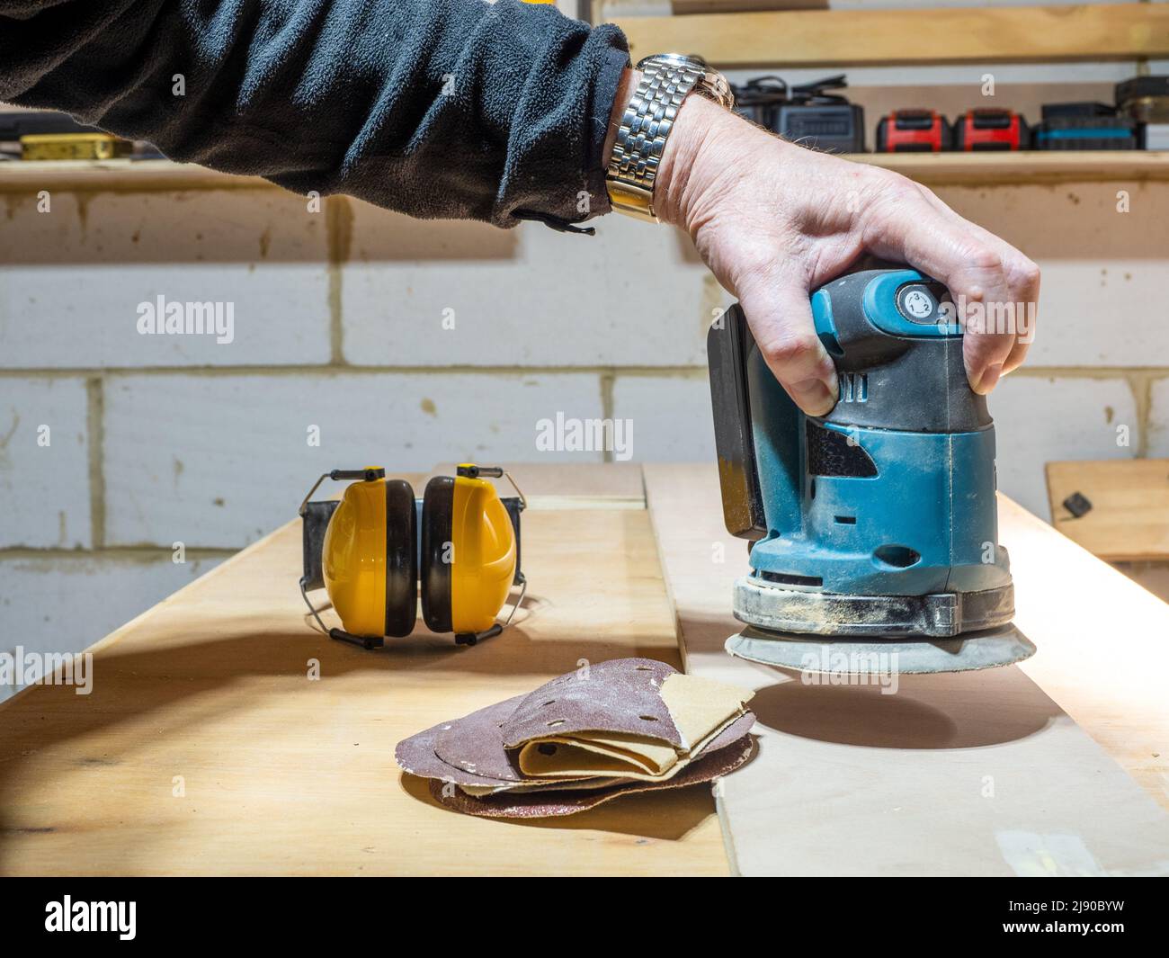 carpenter preparing a plywood board using Cordless battery operated orbital sander with ear defenders  in the background Stock Photo