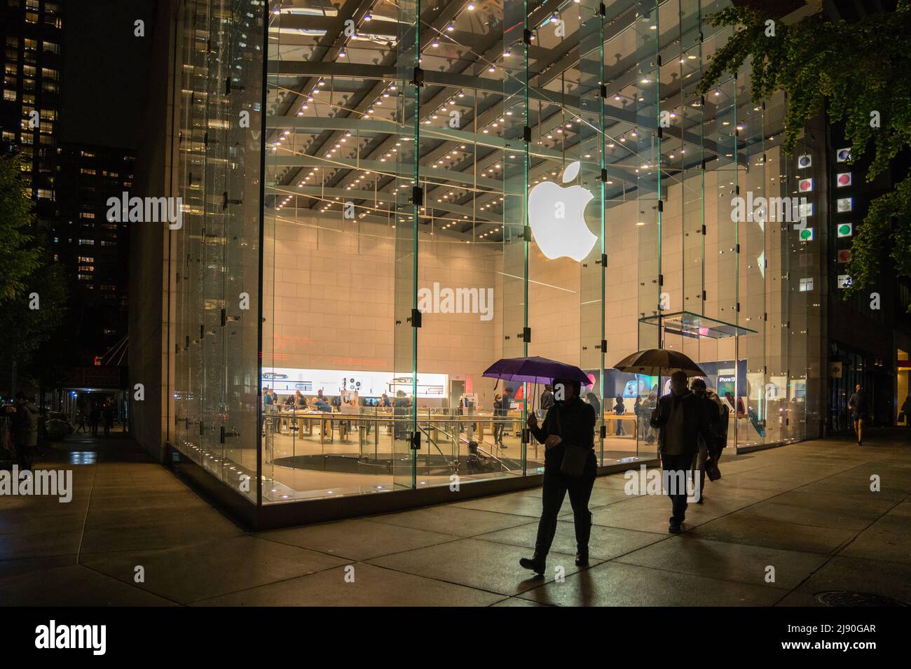 New York, NY, US-October 26, 2021: People with umbrellas walk by Apple computer store on a rainy night. Stock Photo