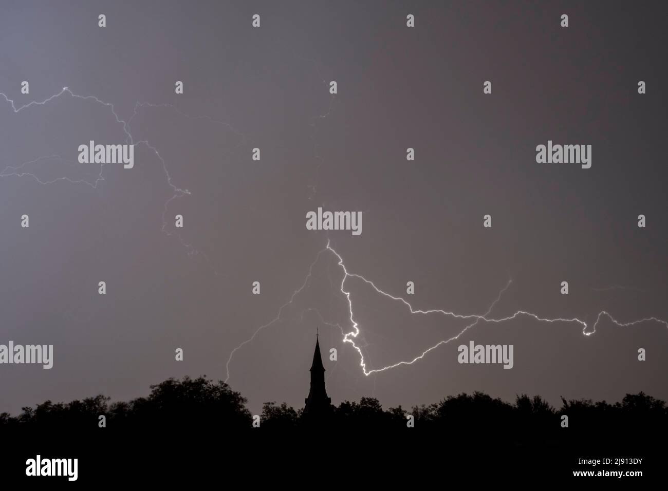 Rain falling and stroke of forked lightning during thunderstorm at night over church tower and trees Stock Photo
