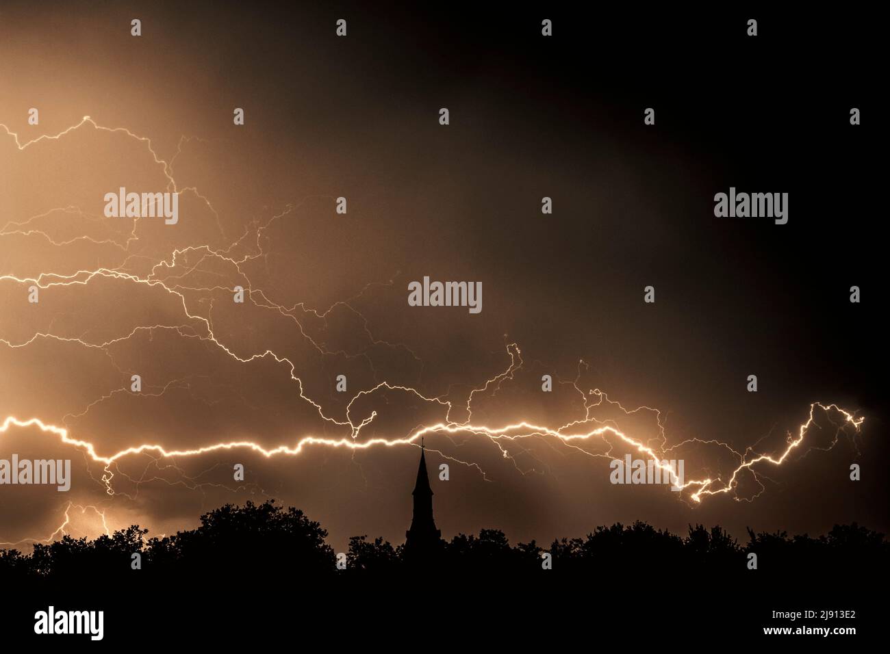 Rain falling and strokes of forked lightnings during thunderstorm at night over church tower and trees Stock Photo