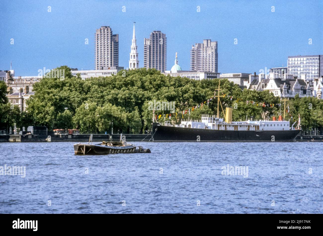 1982 archive image of London skyline seen across River Thames from South Bank - showing towers of the Barbican, spire of St Bride's church and dome of the Old Bailey.  The boat moored on the north bank is HMS Chrysanthemum, an Anchusa class sloop of 1917, scrapped in 1995. Stock Photo