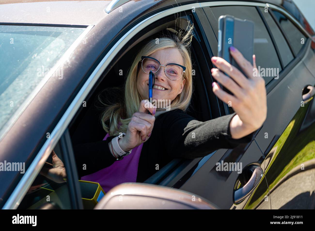 middle-aged blonde woman in glasses making selfie with a smartphone through an open car window Stock Photo