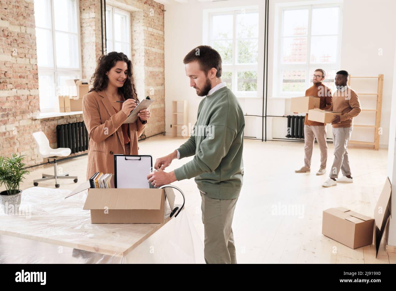 Smiling young woman checking list while her colleague unpacking moving box of documents in new office Stock Photo