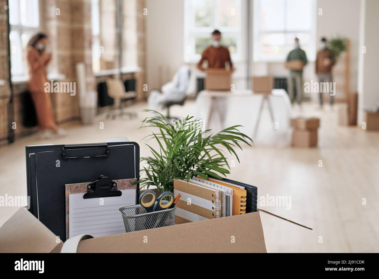 Focus on open cardboard box with office supplies, clipboards and plant against people moving into new office in background Stock Photo