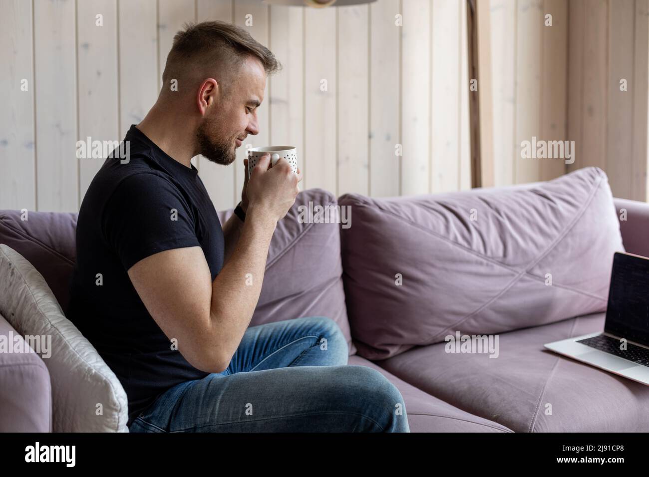 Work at home online. Young man with short stylish haircut in black T-shirt and jeans is sitting on cozy sofa and enjoying a cup of hot coffee, pre Stock Photo