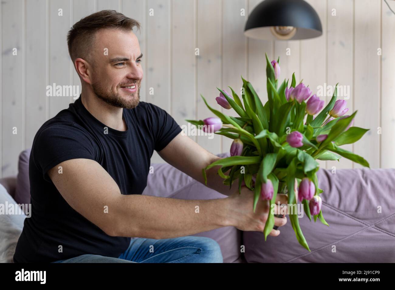 Gute smiling man in black T-shirt and jeans is sitting at home on sofa with bouquet of tulips and preparing surprise for his girlfriend. Preparation Stock Photo