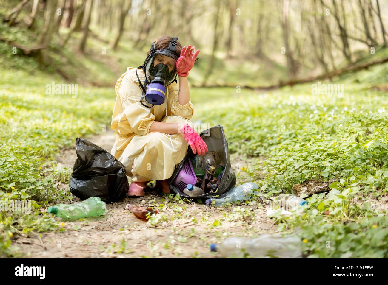 Woman in gas mask and protective clothes collecting scattered plastic garbage in the woods Stock Photo