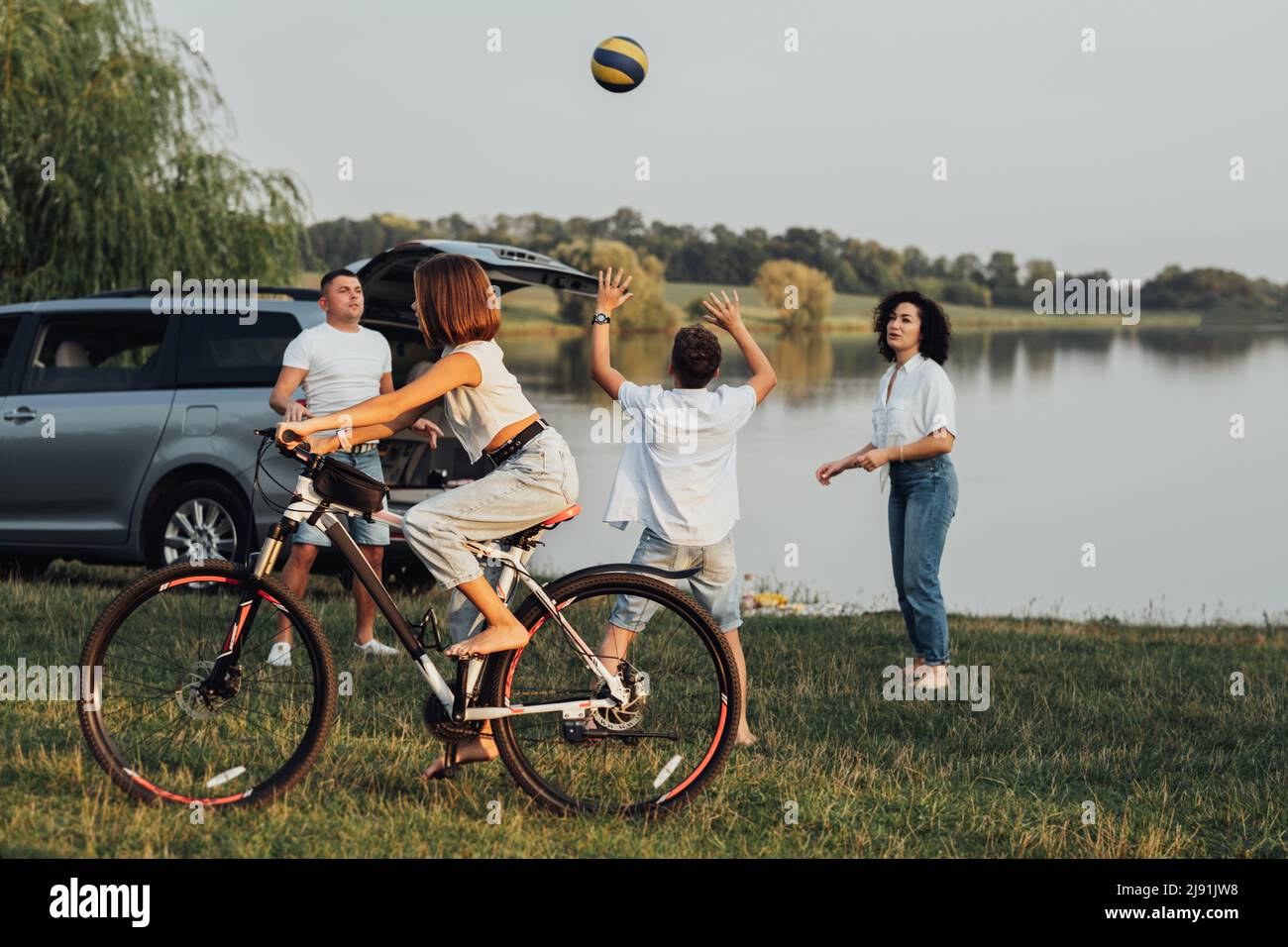 Teenage Girl Riding Bicycle While Mother and Father Playing Volleyball with Her Brother Outdoors by the Lake, Happy Four Members Family Enjoying Stock Photo
