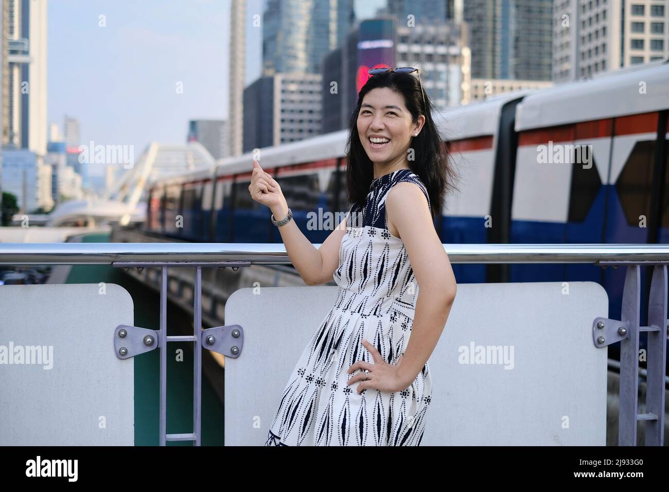 An Asian woman is standing on a platform at a sky train station, feeling happy and smiling with the incoming train arriving in the background. Stock Photo