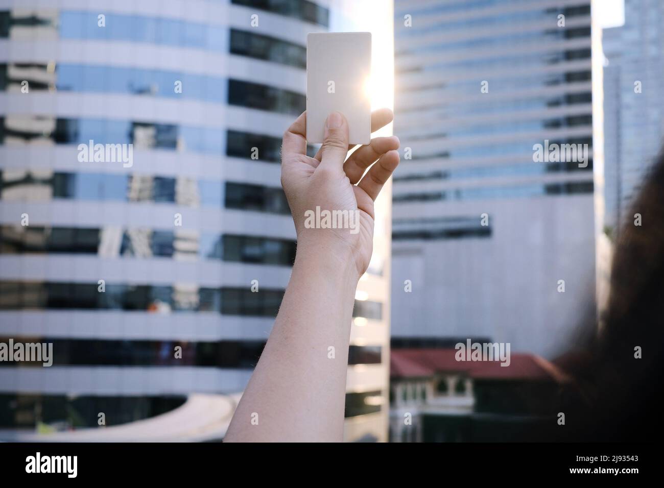 A hand holding up a white blank credit card with sun light shining through on the side and high-rise buildings in the background. Stock Photo