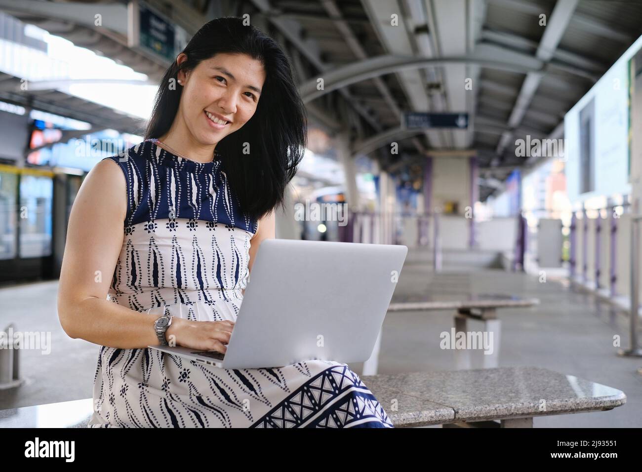 A young business woman is sitting on a bench at a train station or terminal, busy working on her notebook computer while waiting for her train to arri Stock Photo