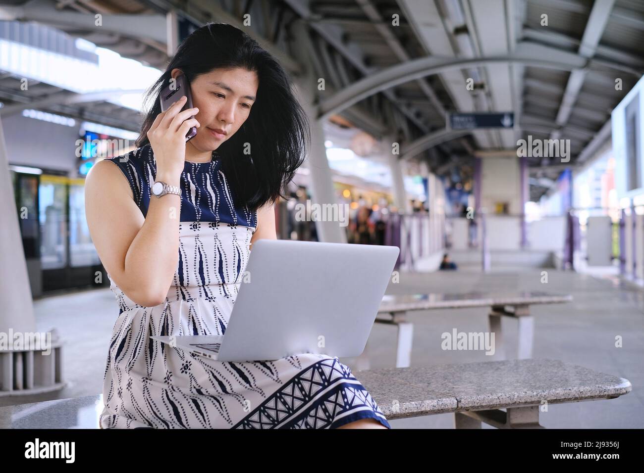 A young business woman is sitting on a bench at a train station or terminal, busy working on her computer and talking on her mobile phone at the same Stock Photo