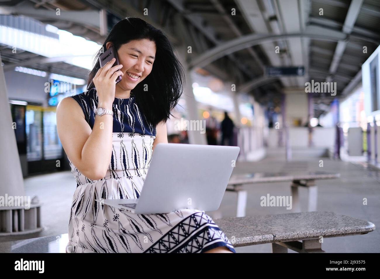 A young business woman is sitting on a bench at a train station or terminal, busy working on her computer and talking on her mobile phone at the same Stock Photo