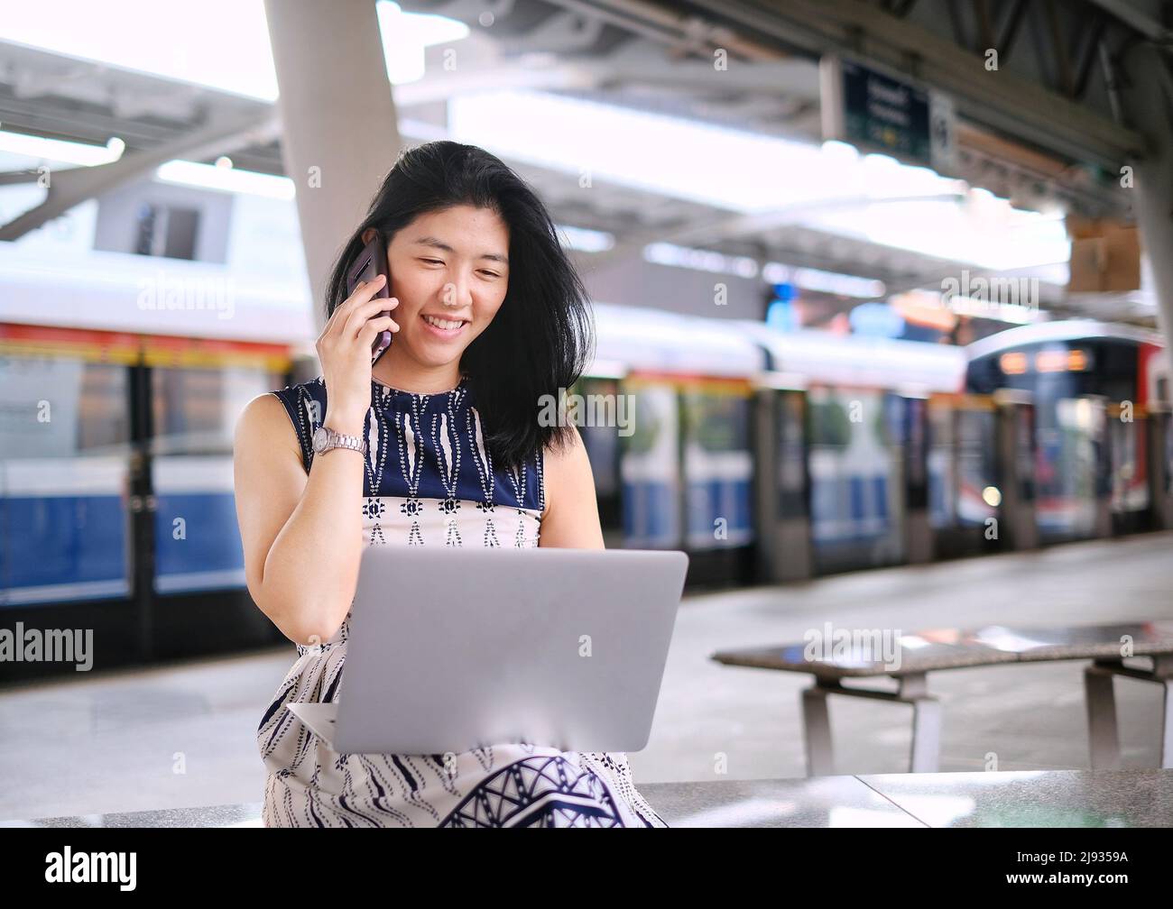 A young business woman is sitting on a bench at a train station or terminal, busy working on her computer and talking on her mobile phone at the same Stock Photo