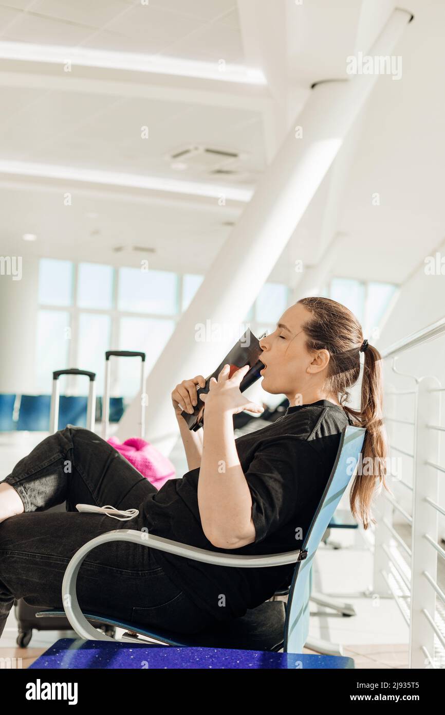 Tired woman sitting with closed eyes on chair in transit lounge of airport, waving with magazine, suffering from heat. Stock Photo