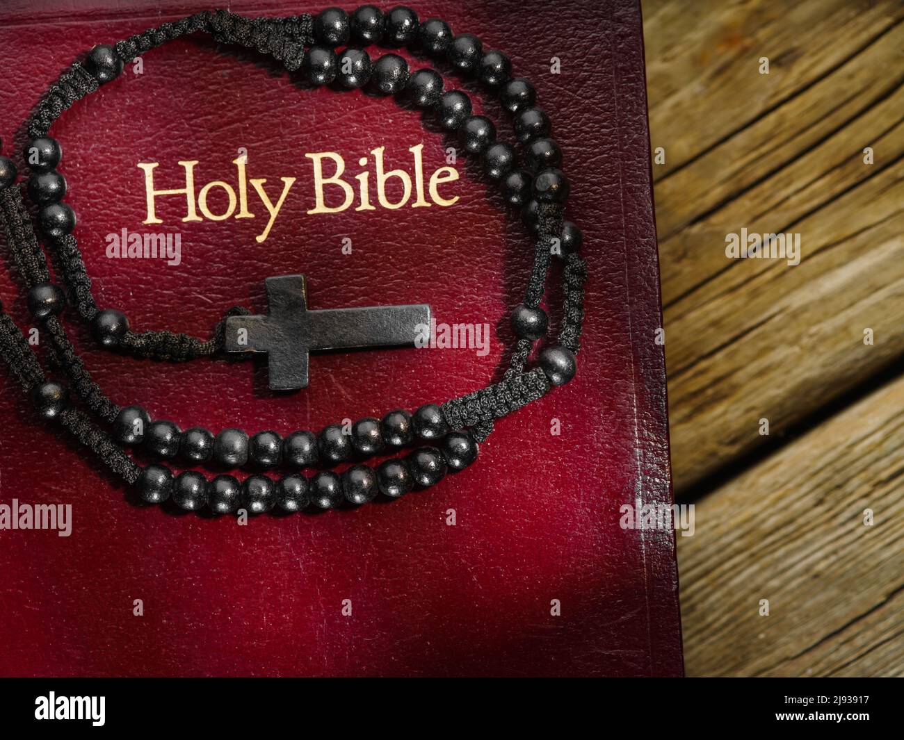 On a simple wooden table lies a Bible and a rosary crucifix. Close-up. There are no people in the photo. Solar lighting. Religious symbols, holy manus Stock Photo