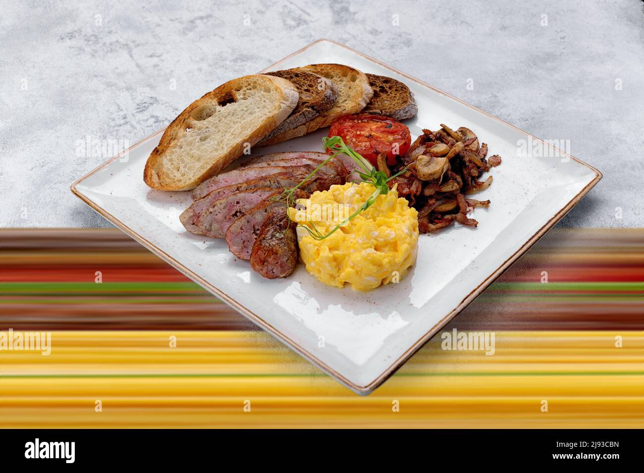 Irish breakfast on a plate. Sausage, scrambled eggs, mushrooms, bacon, ciabatta toast. On a mixed background Stock Photo