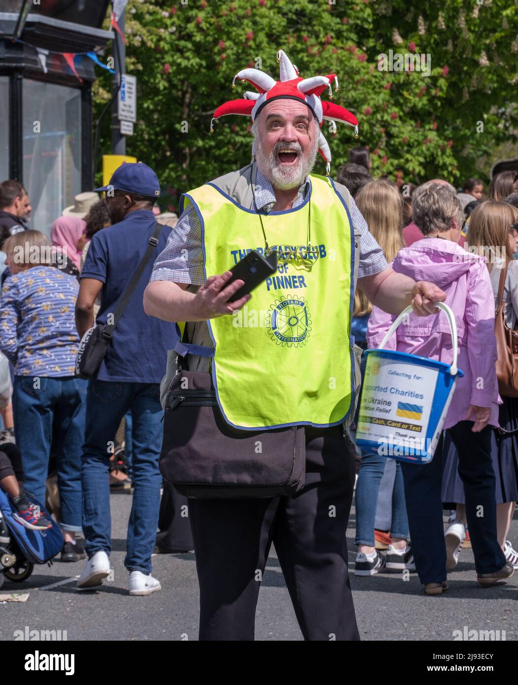 Man wears a Rotary Club of Pinner steward vest & jester hat & holds charity collection bucket for Ukraine, St George’s Day Celebration. Pinner, London Stock Photo