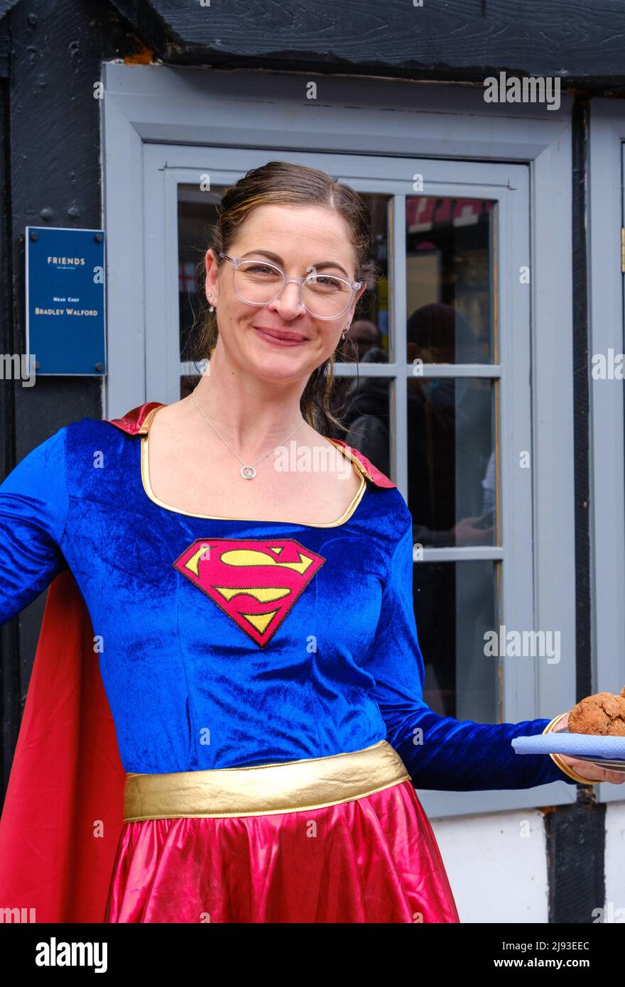 Staff member from Friends Restaurant dressed in Superwoman fancy dress smiles & holds tray of food at 2022 St George’s Day Celebration, Pinner, London Stock Photo