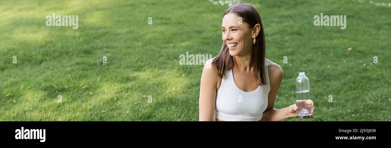 happy woman in white sports bra holding sports bottle and looking away outdoors, banner Stock Photo