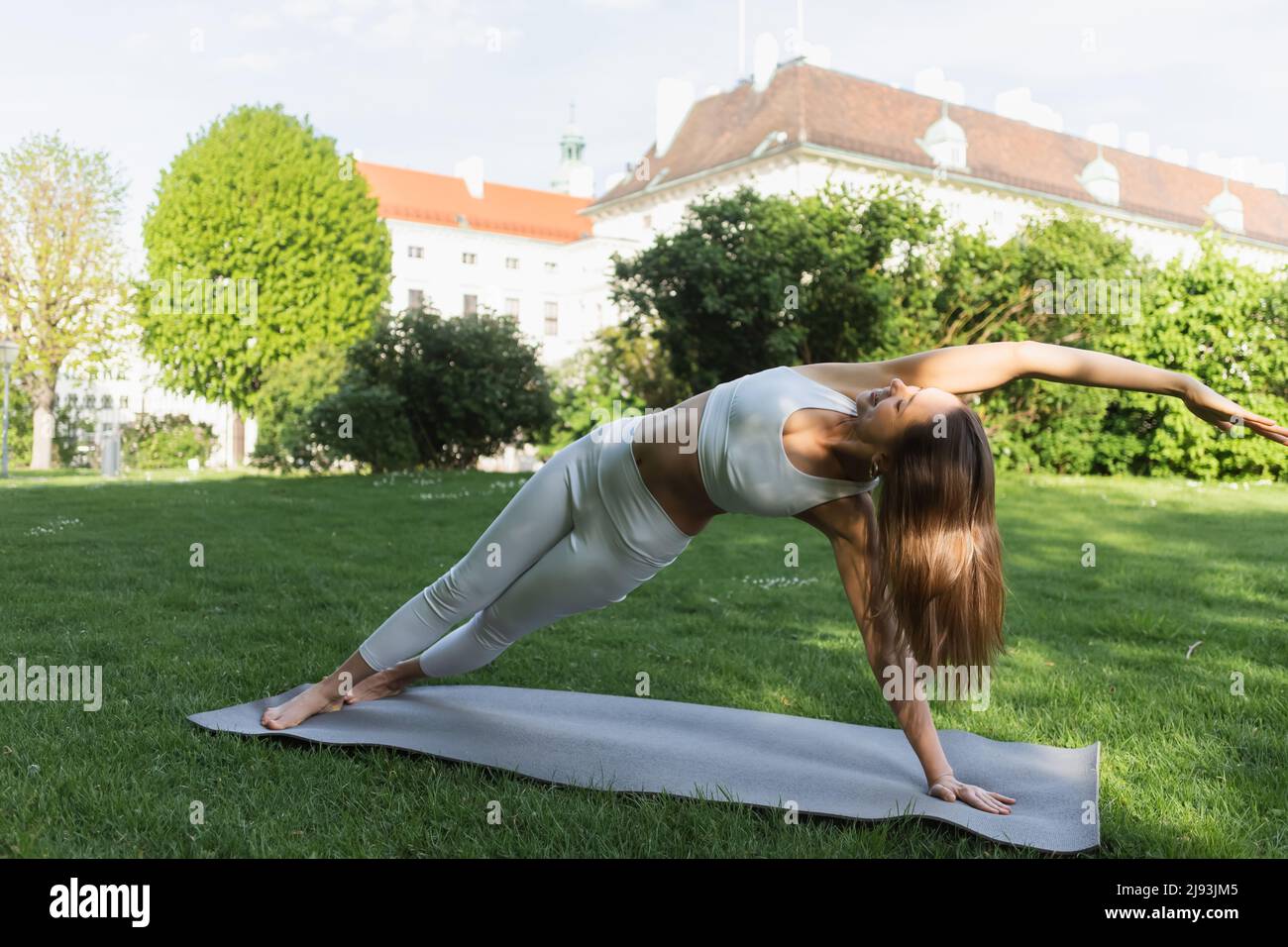 full length of slim woman practicing yoga in side plank pose on yoga mat and green lawn Stock Photo