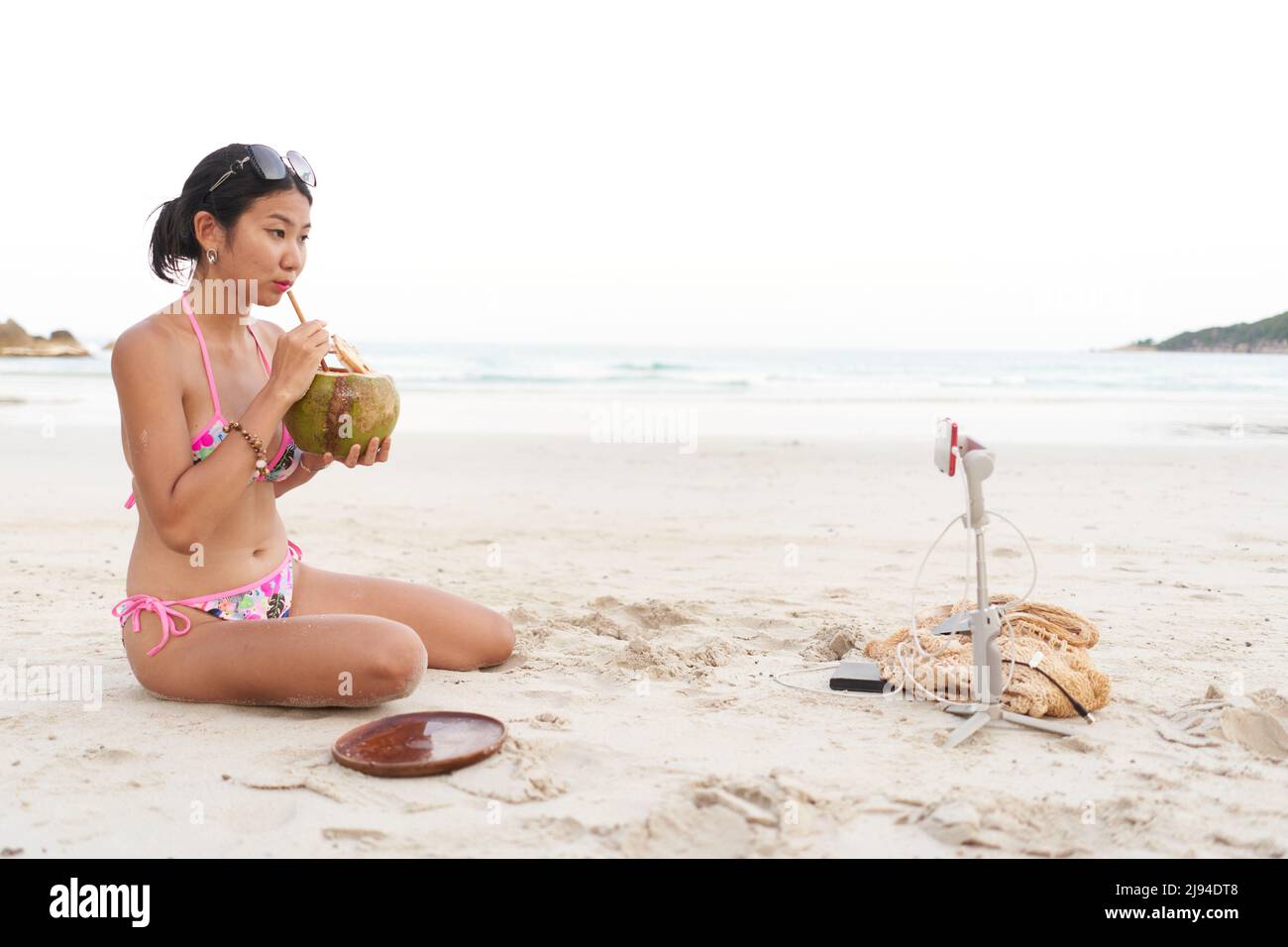Influencer drinking a coconut juice while doing a live using a mobile on the beach Stock Photo