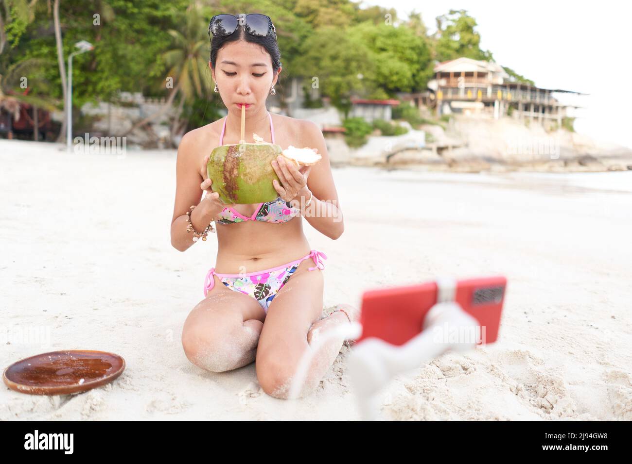 Streamer drinking a coconut juice while doing a live with a mobile on the beach Stock Photo