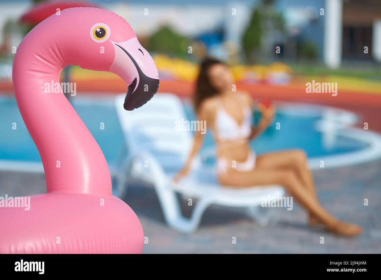 Inflatable pink flamingo ring at swimming pool area outside. Close up view of pink flamingo air mattress with blurred female figure sitting on lounge chair on background. Concept of water activities. Stock Photo