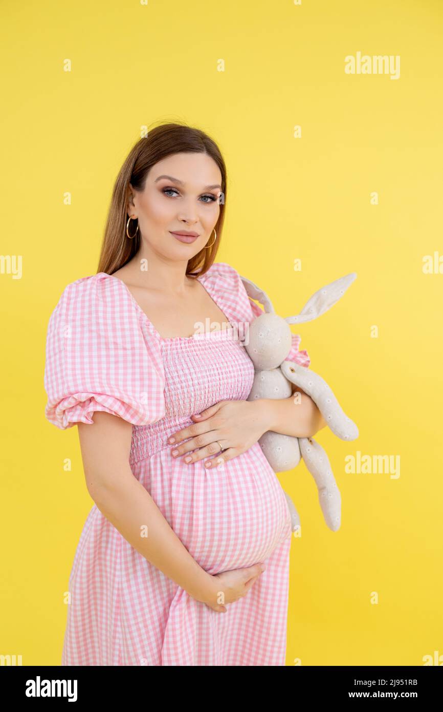 Woman with belly in late pregnancy in casual clothes smile, enjoy and hold soft toy, yellow background. Portrait of young pregnant lady expecting baby Stock Photo