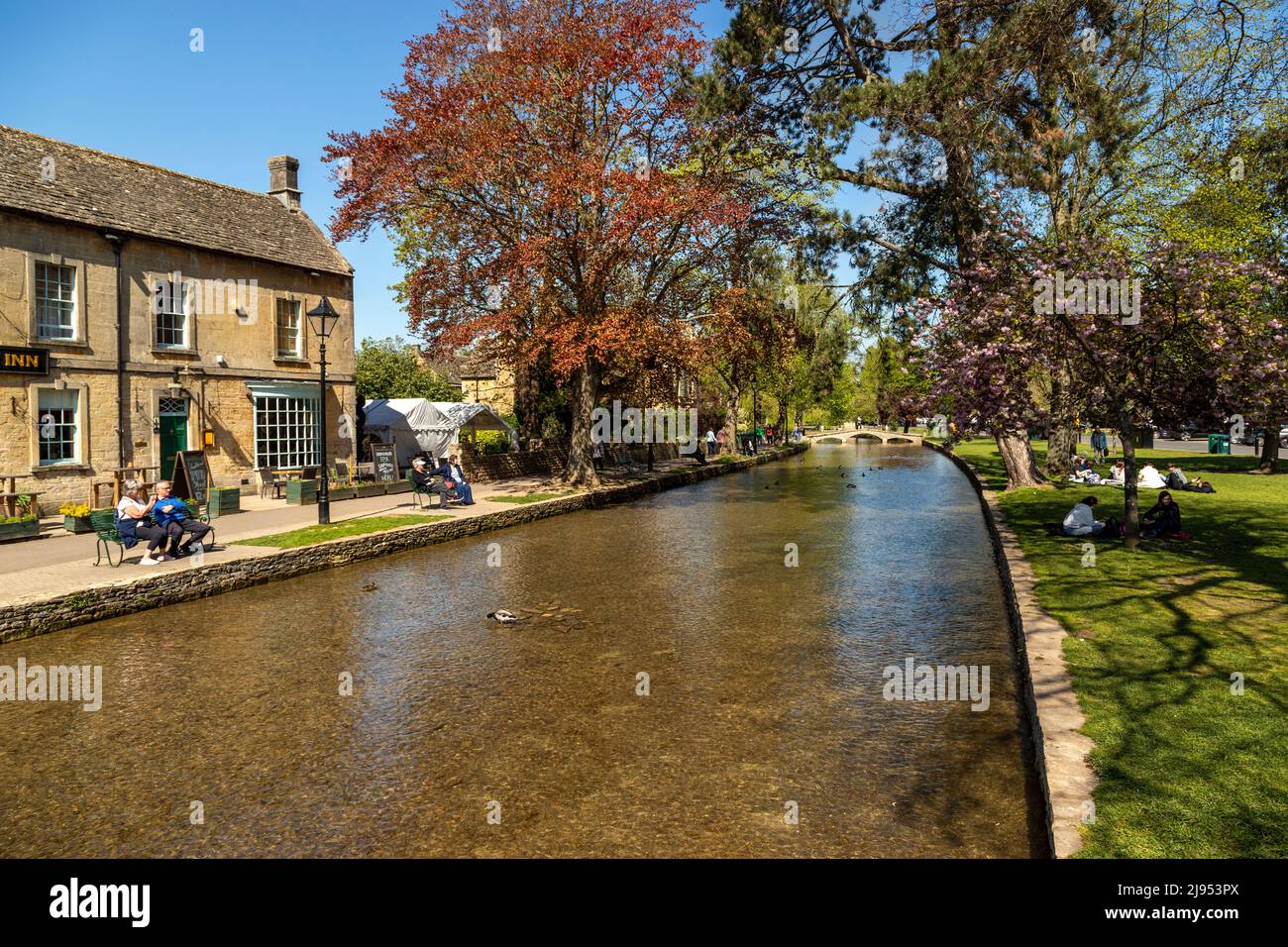 Visitors enjoy a sunny day in Spring in Bourton-on-the-Water along the river Windrush, a popular Cotswolds destination, Gloucestershire, England, UK. Stock Photo