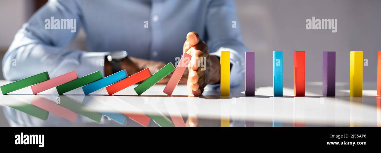 Close-up Of A Businessman Hand Stopping Colorful Dominoes From Falling On Office Desk Stock Photo